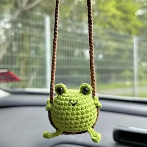 May include: A green crocheted frog hanging from a brown string. The frog has black eyes and a small smile.