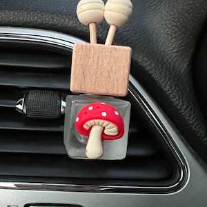 May include: A red and white mushroom shaped car air freshener with a wooden block and two wooden balls on top. The air freshener is hanging from a car's air vent.