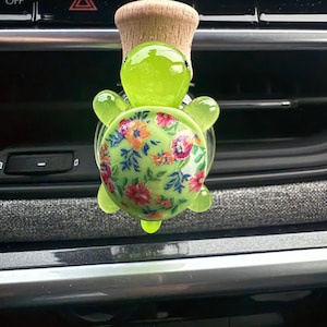 Op de afbeelding: Een groene schildpadvormige autoluchtverfrisser met een bloemenpatroon op zijn schild. De luchtverfrisser is bevestigd aan een houten stok met een houten bal aan de bovenkant.