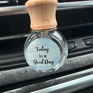 May include: A clear glass car air freshener with a wooden cap. The air freshener has a round sticker with the text "Today is a Good Day" in black script over a light blue watercolor background.