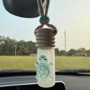 Puede incluir: Un ambientador para coche con un frasco de vidrio esmerilado y una tapa de madera marrón claro. Un dije de tortuga azul claro está adherido a la botella. El ambientador cuelga de una cuerda trenzada con una cuenta turquesa.