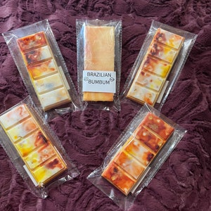 May include: Five wax melts in clear packaging. The wax melts are in various shades of yellow and orange with a marbled effect. The wax melts are labeled "Brazilian Bumbum".