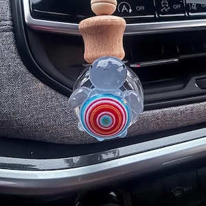 Puede incluir: Un ambientador de coche de vidrio azul en forma de tortuga con un diseño de espiral colorido. El ambientador está unido a un palo de madera con una cuenta de madera en la parte superior.