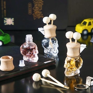 May include: Three glass skull-shaped bottles with wooden lids and decorative sticks. The bottles are filled with a clear liquid and are arranged on a dark surface.