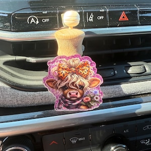 Puede incluir: Un ambientador decorativo para coche con un diseño de vaca de las Tierras Altas. La vaca tiene pelo claro, un lazo con estampado de leopardo y un ramo de flores. El ambientador está unido a una base de madera con una parte superior blanca.