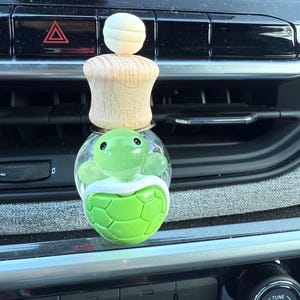 May include: A car air freshener featuring a green turtle design. The turtle is inside a clear glass container, with a wooden base and a small, round wooden top. The air freshener is designed to clip onto a car's air vent.