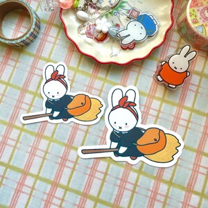 May include: A selection of stickers and acrylic charms showcasing a white rabbit character. The rabbit is shown in different poses, such as riding a broom and wearing an orange outfit. Decorative tape and a small dish are also visible.