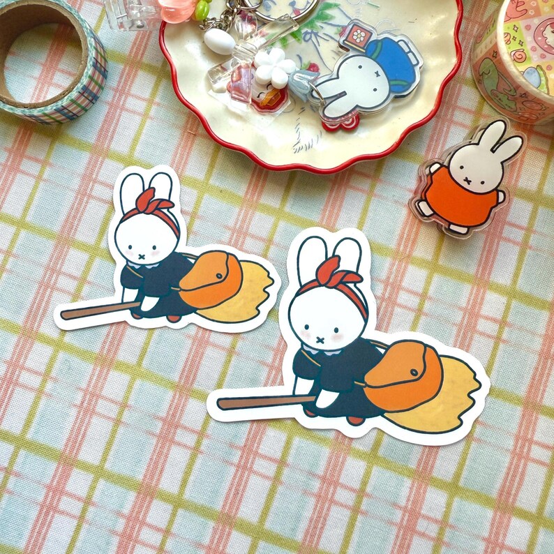 May include: A selection of stickers and acrylic charms showcasing a white rabbit character. The rabbit is shown in different poses, such as riding a broom and wearing an orange outfit. Decorative tape and a small dish are also visible.