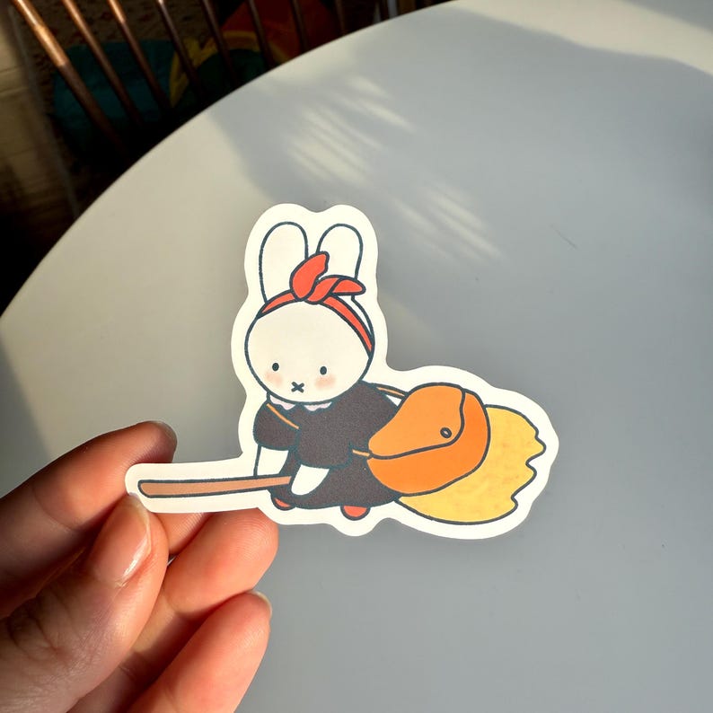 May include: A sticker of a cartoon bunny in a black outfit and red headband, riding a broom. The bunny has an orange bag and the broom has a yellow brush. The sticker is held by a hand.