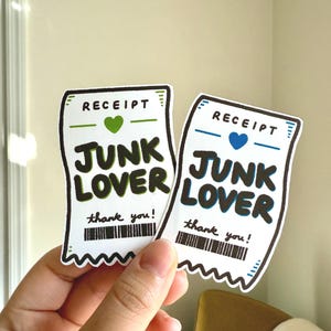 May include: Two white stickers in the shape of receipts, held by a hand. The stickers have the word "RECEIPT" at the top, a heart, and the words "JUNK LOVER" in bold letters. Below is a barcode and the words "thank you!"