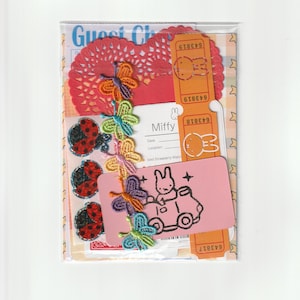 Puede incluir: Un conjunto de artículos de papelería que incluye un tapete de papel rojo, boletos de rifa naranjas con iconos de conejo Miffy, tres pegatinas de mariquitas, una fila de mariposas de tela coloridas y una tarjeta rosa con un conejo Miffy.