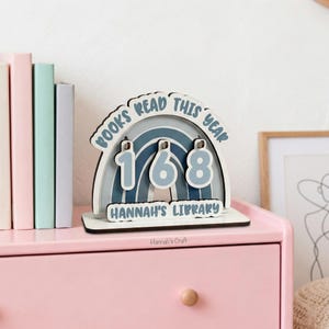 May include: A wooden book counter with a rainbow design, displaying the number "168" in blue. The text "BOOKS READ THIS YEAR" arches above, and "HANNAH'S LIBRARY" is below. The counter sits on a pink surface.