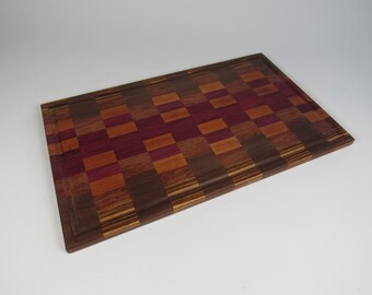 May include: Rectangular wooden cutting board with a checkerboard pattern. The board features a mix of dark brown, reddish-purple, and light brown wood tones. A recessed groove runs along the perimeter.