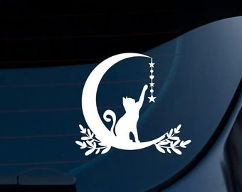 May include: A white vinyl decal applied to a car window featuring a silhouette of a cat sitting on a crescent moon. The cat is reaching up to touch a vertical string of hanging stars and hearts. Decorative leafy sprigs accent the base of the moon.