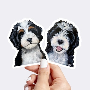 May include: Two black and white doodle dog stickers with a white background. The stickers are painted in a realistic style and feature the dogs' faces. The text "Teresa Curella Art" is visible on the bottom right sticker.