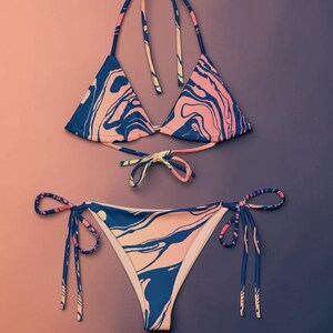 Può includere: Un bikini a due pezzi con un motivo a vortice simile al marmo nei toni del blu navy e del pesca tenue. Il top presenta coppe triangolari con laccetti da annodare al collo e sulla schiena, mentre lo slip è dotato di laccetti laterali regolabili.