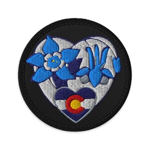 May include: Embroidered patch with a heart design, blue flowers, and the Colorado state flag. The patch has a black border and is suitable for fabric items. The flowers have blue and white petals. Ideal for jackets or bags.