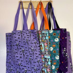 May include: A collection of five fabric tote bags hanging on a wooden rack. The bags feature various patterns inspired by The Nightmare Before Christmas, including Jack Skellington, Sally, bats, pumpkins, and colorful stained glass motifs.