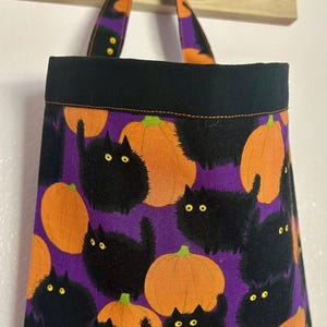 May include: A Halloween-themed tote bag with a black top and handles. The bag's body features a purple background with orange pumpkins and black cat illustrations. The handles have an orange and purple pattern.
