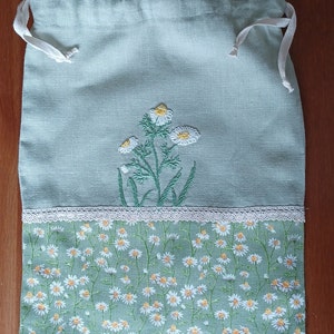 May include: A light blue drawstring bag with a white lace trim. The bag is decorated with embroidered white daisies and a green stem. The bottom of the bag is printed with a pattern of white daisies on a green background.