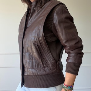 May include: A dark brown leather jacket featuring a quilted square pattern on the front and sleeves. The jacket includes a front zip closure, a collar, and dark brown ribbed knit cuffs and a ribbed hem. The side panels are made of ribbed fabric.