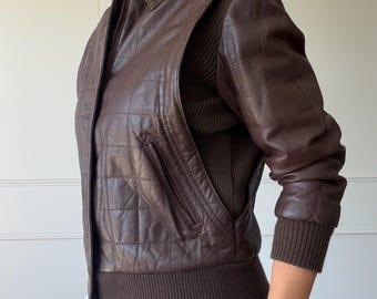 Può includere: Una giacca in pelle marrone scuro con un motivo trapuntato a quadrati sul davanti e sulle maniche. La giacca presenta una chiusura frontale con zip, un colletto e polsini e orlo in maglia a coste di colore marrone scuro sui fianchi.