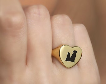 Op de afbeelding: Een goudkleurige hartvormige ring met een zwart silhouet van een kat en een hond. De ring wordt aan een vinger gedragen. De ring is een sieraad.
