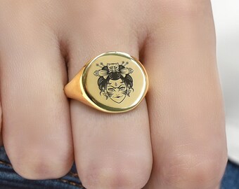 May include: A gold signet ring featuring a black and white image of a woman's face with flowers in her hair.