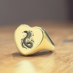 Puede incluir: Un anillo de oro en forma de corazón con un diseño de serpiente en blanco y negro grabado en la parte delantera. La serpiente está enroscada y tiene un patrón detallado.