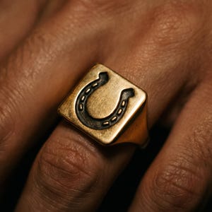 May include: A gold-toned signet ring featuring a square face with a recessed, dark-colored horseshoe emblem engraved in the center. The metal surface has a brushed, matte finish, and the band tapers slightly toward the back of the finger.