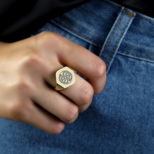 May include: A gold signet ring with a black stone design. The ring is worn on a person's finger.