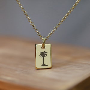 Puede incluir: Un collar de cadena dorada con un colgante rectangular que presenta una silueta de una palmera.