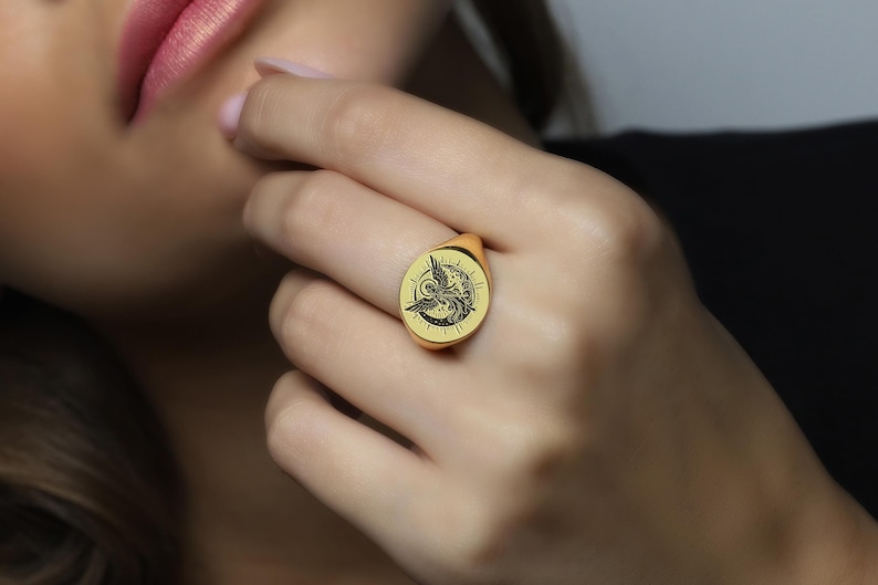 May include: A gold signet ring with an engraved image of an angel with wings spread. The ring is worn on the index finger of a hand.