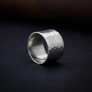 A silver ring with a textured surface sits on a dark background.