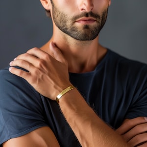 May include: A gold-toned bracelet with an engraved inscription. The bracelet is worn on the left wrist of a person wearing a dark blue t-shirt.