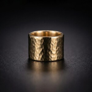 A shiny gold ring with a textured surface sits on a dark surface, creating a striking contrast.