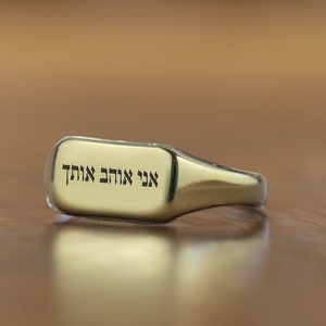 Puede incluir: Un anillo de sello de oro con una parte superior rectangular que presenta texto en hebreo que dice "Ani Ma'ahech Odcha", que se traduce como "Yo estoy contigo siempre".
