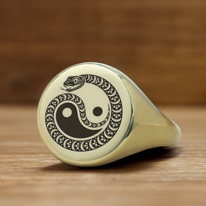 May include: A gold signet ring with a black and white yin yang symbol and a snake wrapped around it.