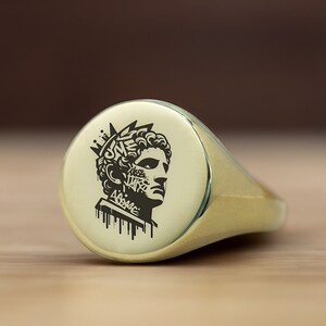May include: A gold signet ring with a black and white engraved image of a person's head with a crown and graffiti-style text. The text reads "ONE WAY" and "AGAPE".