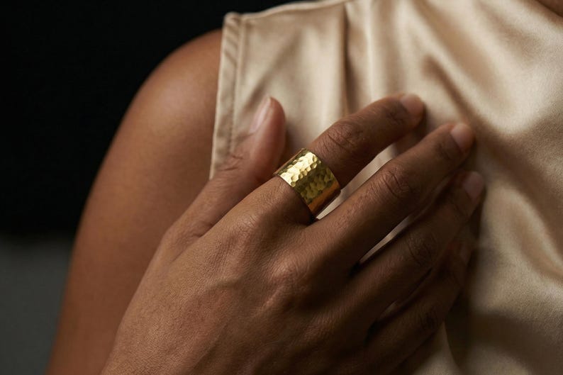 A close-up of a person's hand wearing a gold ring, with a beige fabric sleeve visible.