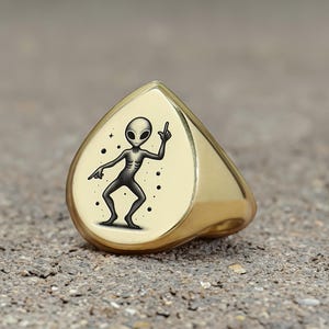 May include: A gold ring with a teardrop-shaped design featuring a black and white illustration of an alien dancing. The alien has a large head and big eyes.