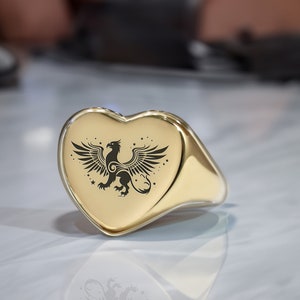 Könnte beinhalten: Ein goldener Herzring mit einem gravierten schwarzen Greifendesign auf der Vorderseite.