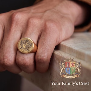 May include: A gold signet ring featuring a circular face engraved with a detailed heraldic crest and the text Honor & Strength. The ring has a polished metallic finish and a smooth, rounded band design, showcasing a classic and substantial appearance.