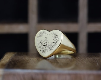 May include: A gold heart-shaped ring with an engraved design of two birds perched on a branch with flowers and leaves.