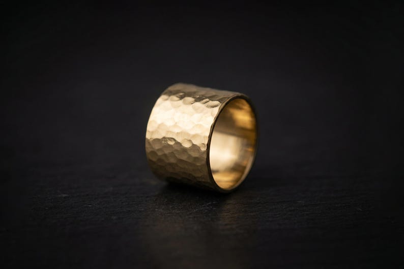 A shiny gold ring with a textured surface sits on a dark surface, creating a striking contrast.
