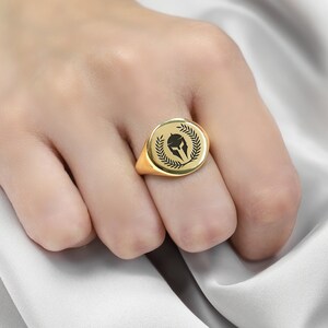 Puede incluir: Un anillo de sello de oro con una imagen en blanco y negro de un casco espartano dentro de una corona de laurel.