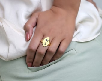 May include: Gold ring with an oval face displaying a black palm tree motif. The ring is on a hand, with a white blouse and pale green trousers visible in the background.