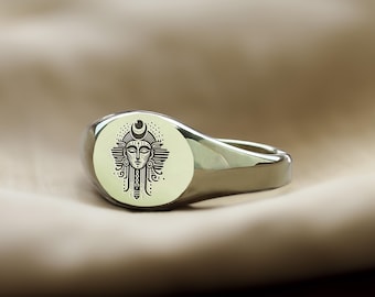 May include: A silver signet ring with an oval-shaped top featuring a detailed engraved design of a woman's face with a crescent moon above her head.