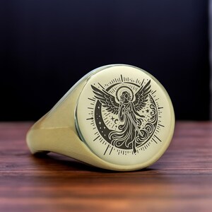 May include: A gold signet ring with a detailed engraving of an angel with wings spread, surrounded by stars and a crescent moon.