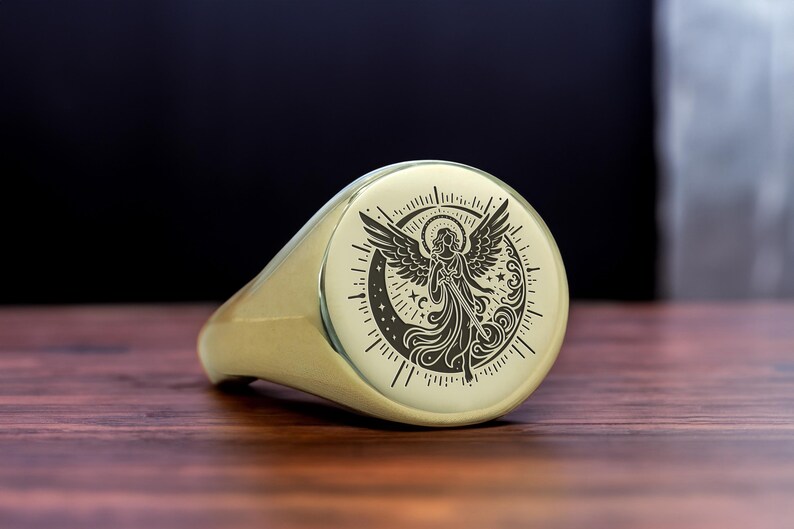 May include: A gold signet ring with a detailed engraving of an angel with wings spread, surrounded by stars and a crescent moon.